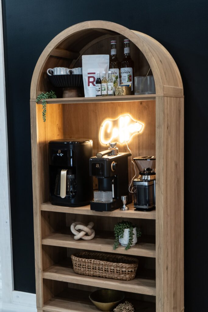 A wooden shelf holds coffee machines, bottles, cups, and decorative plants, with a neon “Café” sign glowing beside the machines against a dark wall background.