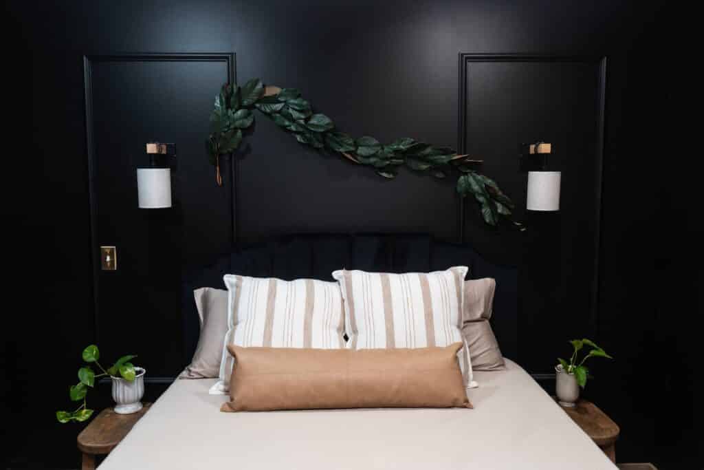 A modern bedroom with a dark wall, a bed with white and beige pillows, a long tan cushion, and a curved green leafy garland above the headboard—perfect for relaxing in one of Ohio’s luxury cabins. Small potted plants sit on wooden nightstands on each side.