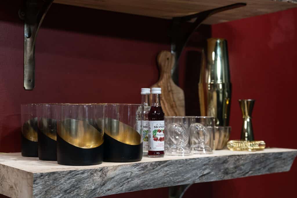 A wooden shelf in luxury cabins holds black and gold glasses, clear glasses, two bottles of syrup, a gold cocktail shaker, a jigger, and a muddler against a dark red wall—perfect for beautiful vacations.