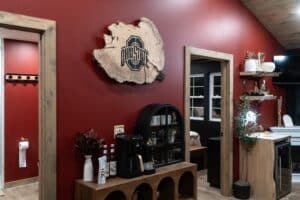 A coffee station with a coffee maker, mugs, and bottles sits against a red wall featuring a large Ohio State University logo plaque—adding collegiate charm to luxury cabins in Laureville. Adjacent rooms and rustic decor are visible in the background.