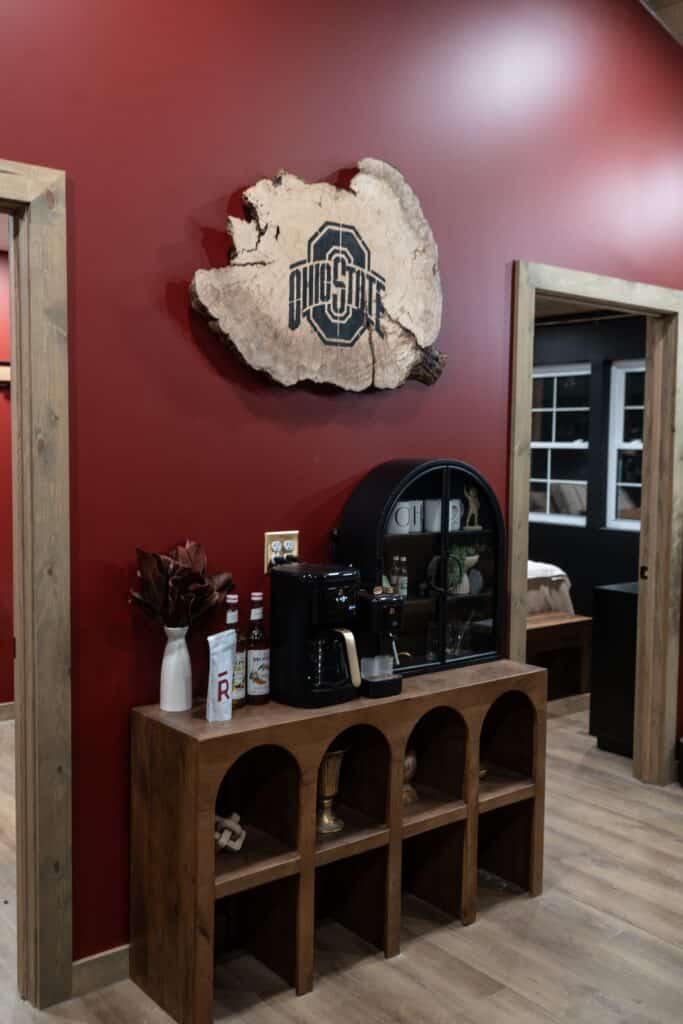 A wooden console table with a coffee maker, mugs, and bottles sits against a red wall in this inviting space. Above, a large wooden Ohio State University logo sign is mounted—perfect for luxury cabin stays with wood trim and hardwood floors.