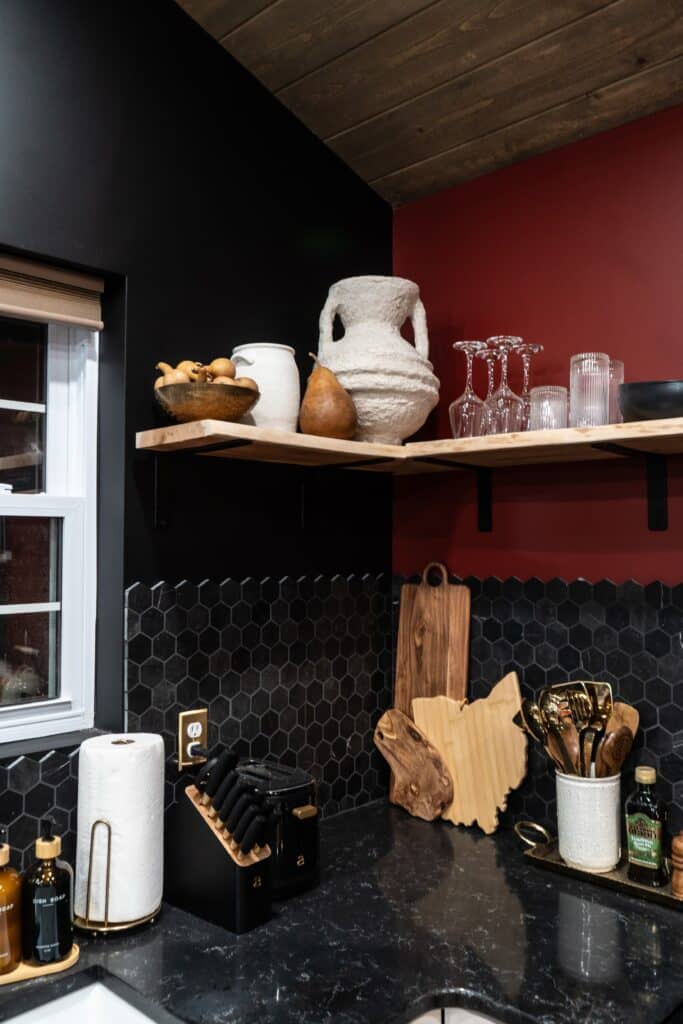 A modern kitchen corner with black hexagon tile backsplash, wooden shelves holding vases, glasses, and bowls—bringing a touch of home sweet Ohio charm to your space—plus a countertop with utensils, cutting boards, knife block, paper towels, and oil bottles.