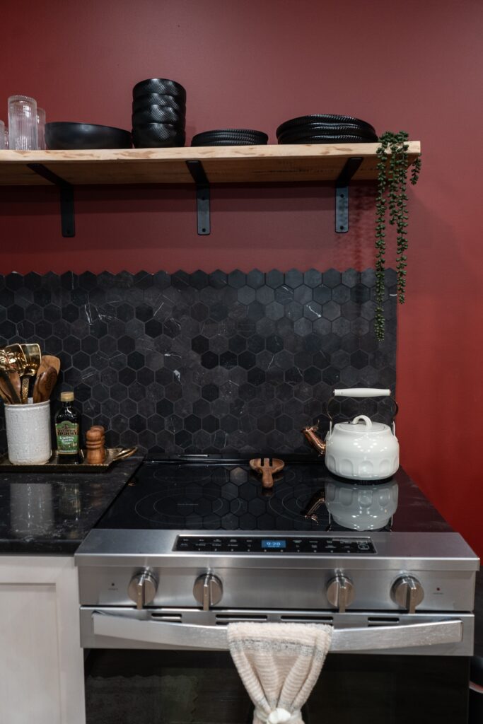 A modern kitchen with a stainless steel stove, white kettle, black hexagonal tile backsplash, wooden utensils, and a shelf with black dishes and a hanging plant—perfect for those seeking luxury cabin stays or that home sweet Ohio charm.