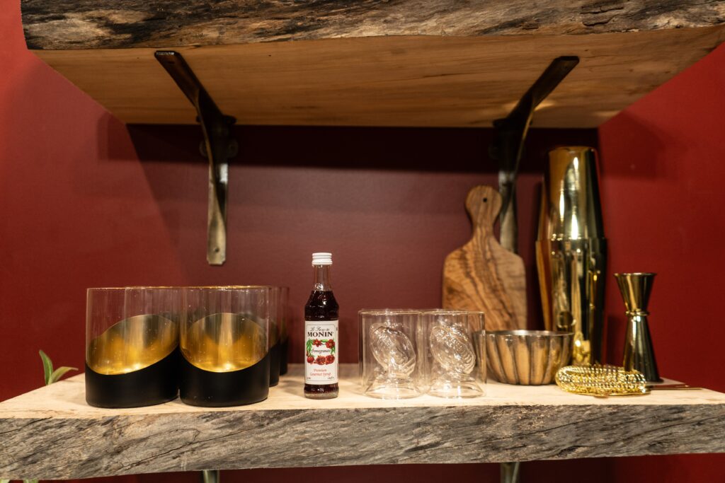 A wooden shelf holds cocktail glasses, a small bottle of Monin raspberry syrup, two textured glasses, a cutting board, shaker, jigger, and strainer—perfect touches for relaxing nights in luxury cabin stays or your home sweet Ohio.