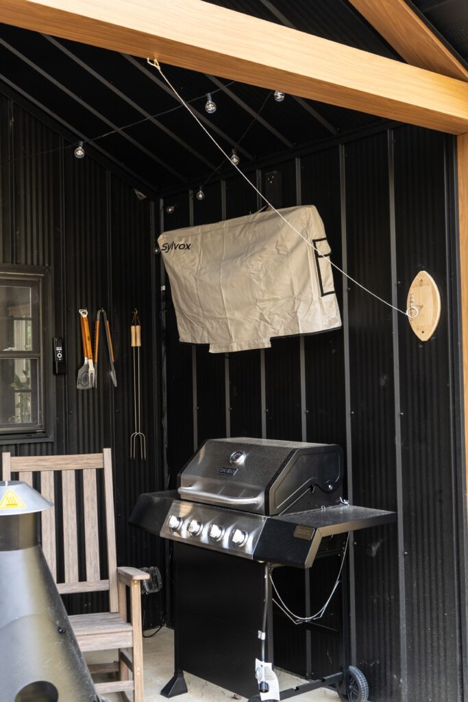 A covered outdoor grilling area at one of our luxury cabins, featuring a black barbecue grill, hanging grill cover, cooking utensils on hooks, a wooden chair, and string lights above—all set against a dark corrugated wall.