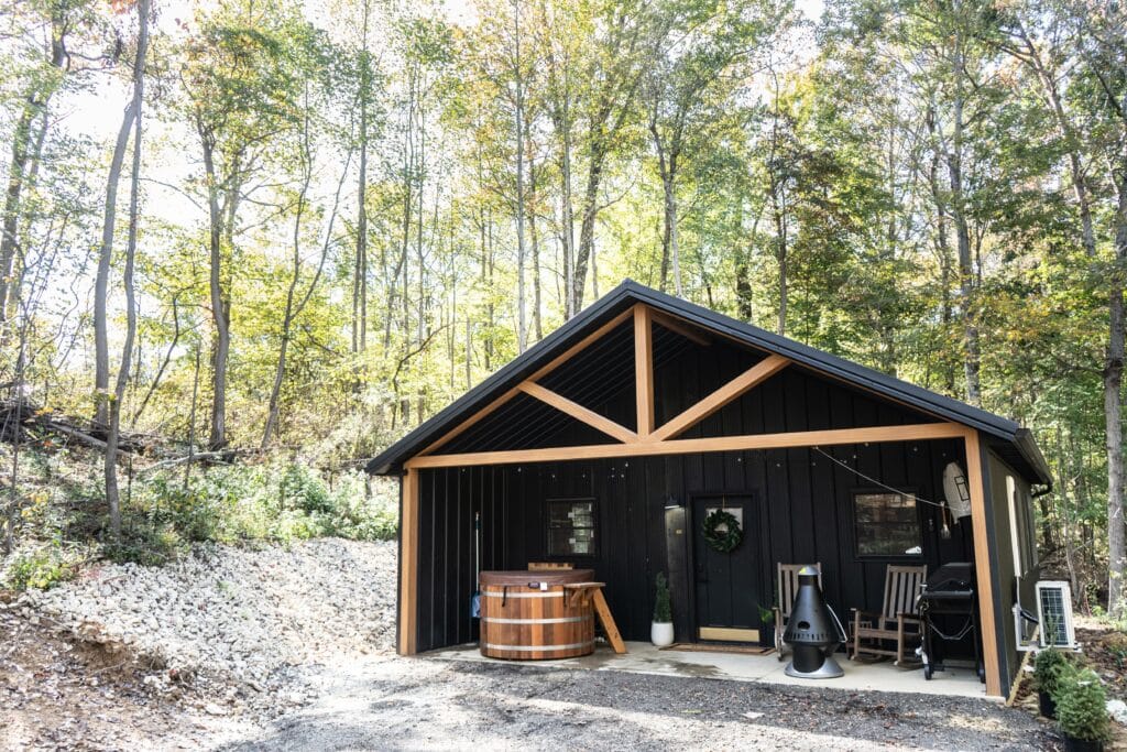 A modern black cabin with wood accents sits in a wooded area. In front, a wooden hot tub, chiminea, rocking chairs, and string lights invite relaxation. Surrounded by green foliage and sunlight—perfect for beautiful vacations or cabins in Laureville.